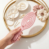 Pink paddle brush held by a hand with pearls and scrunchies on a tray in the background.