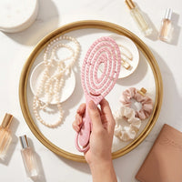 Hand holding a pink hairbrush over a tray with pearls, scrunchies, and perfume bottles.